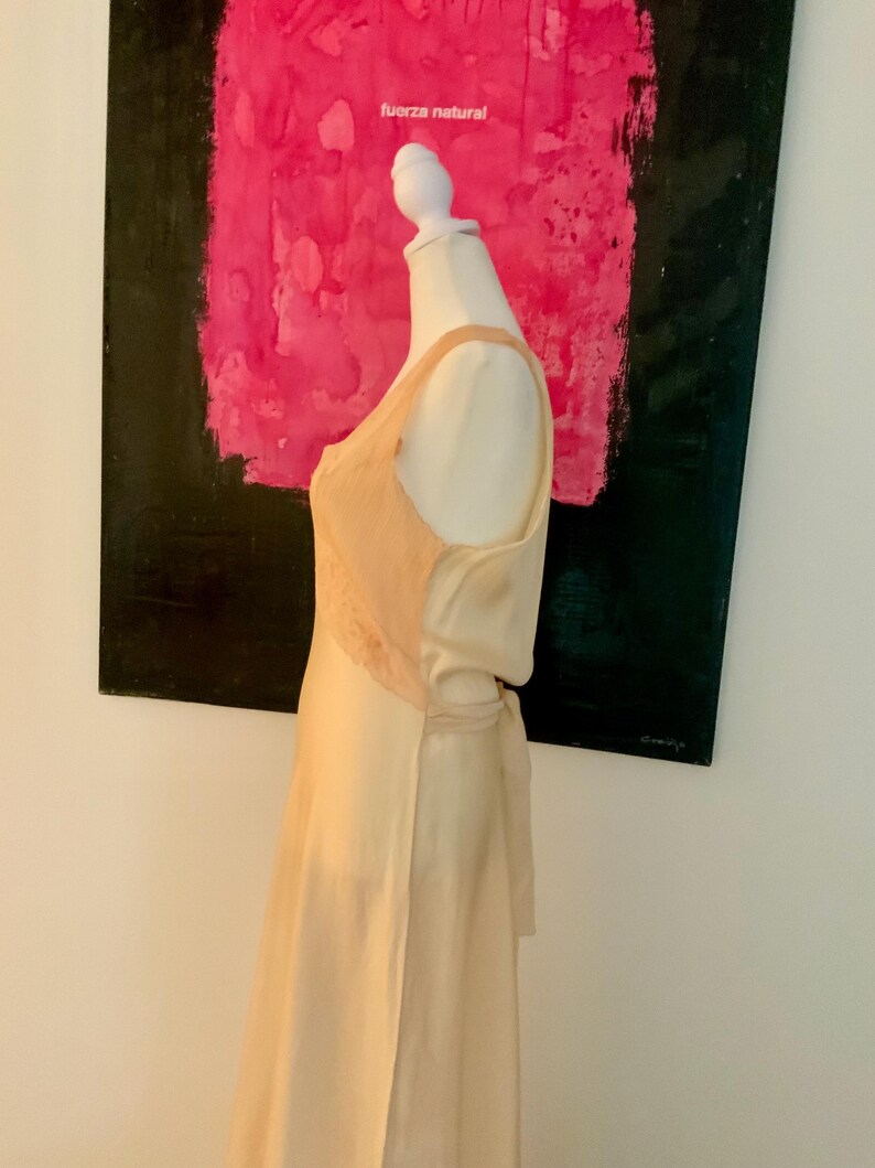 May include: A cream-colored silk dress on a mannequin. The dress features a lace panel on the bodice and a tie at the waist. The background includes a painting with the words "fuerza natural".