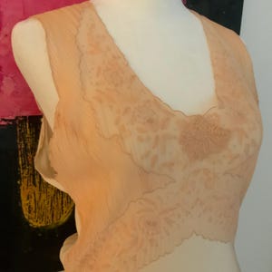 May include: A peach-colored, sleeveless slip with a V-neckline. The slip features a delicate floral lace design across the bodice and a scalloped edge. The fabric appears to be a lightweight, semi-sheer material.