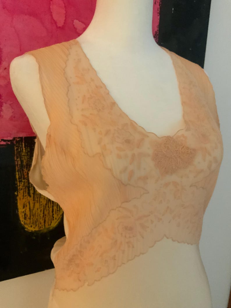 May include: A peach-colored, sleeveless slip with a V-neckline. The slip features a delicate floral lace design across the bodice and a scalloped edge. The fabric appears to be a lightweight, semi-sheer material.