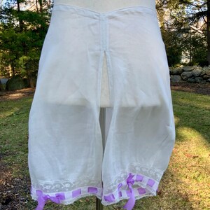 May include: White vintage bloomers with a split front, displayed on a mannequin. The bloomers feature a delicate lace trim along the hemline, accented with purple ribbon bows. The fabric appears sheer and lightweight, suggesting a vintage style.