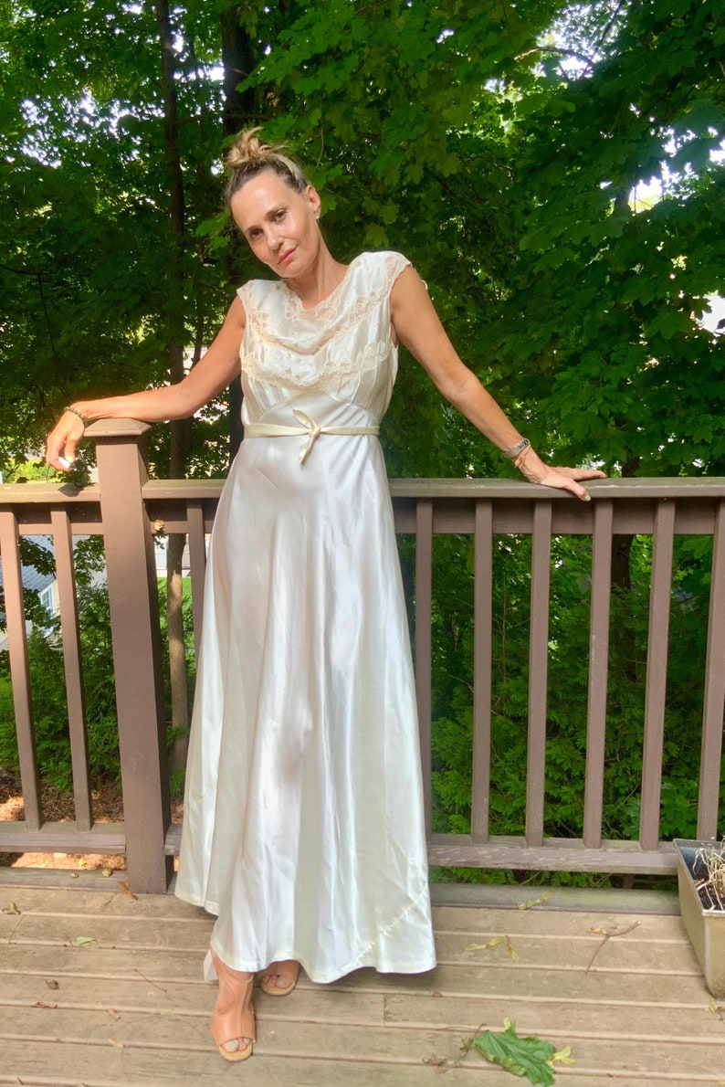 Pode incluir: Um vestido branco de cetim com um corpete de renda e uma faixa de cetim. O vestido &eacute; longo e tem um corpete justo. A modelo est&aacute; em p&eacute; em um deck de madeira com &aacute;rvores no fundo.
