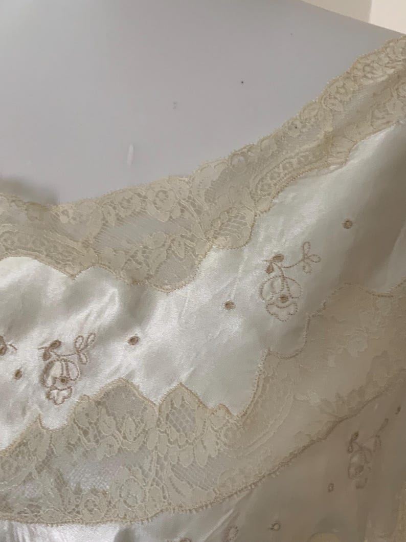 Pode incluir: Uma camisola de cetim branco com renda delicada e detalhes florais bordados.