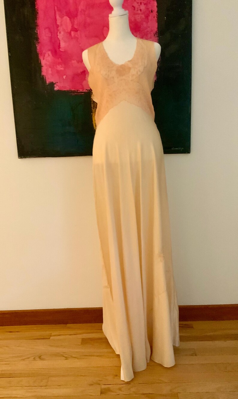 May include: A vintage-style, floor-length peach-colored slip dress on a mannequin. The dress features a delicate lace bodice and a flowing, sheer skirt. The mannequin is positioned in front of a colorful abstract painting.