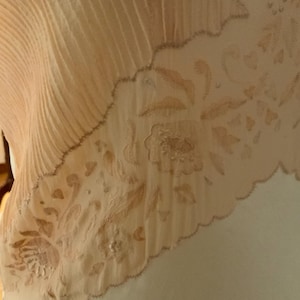 May include: Close-up of a cream-colored textile with a pleated section and a floral embroidered border. The embroidery features detailed flowers and leaves in a soft, muted peach color. The edge has a scalloped design.