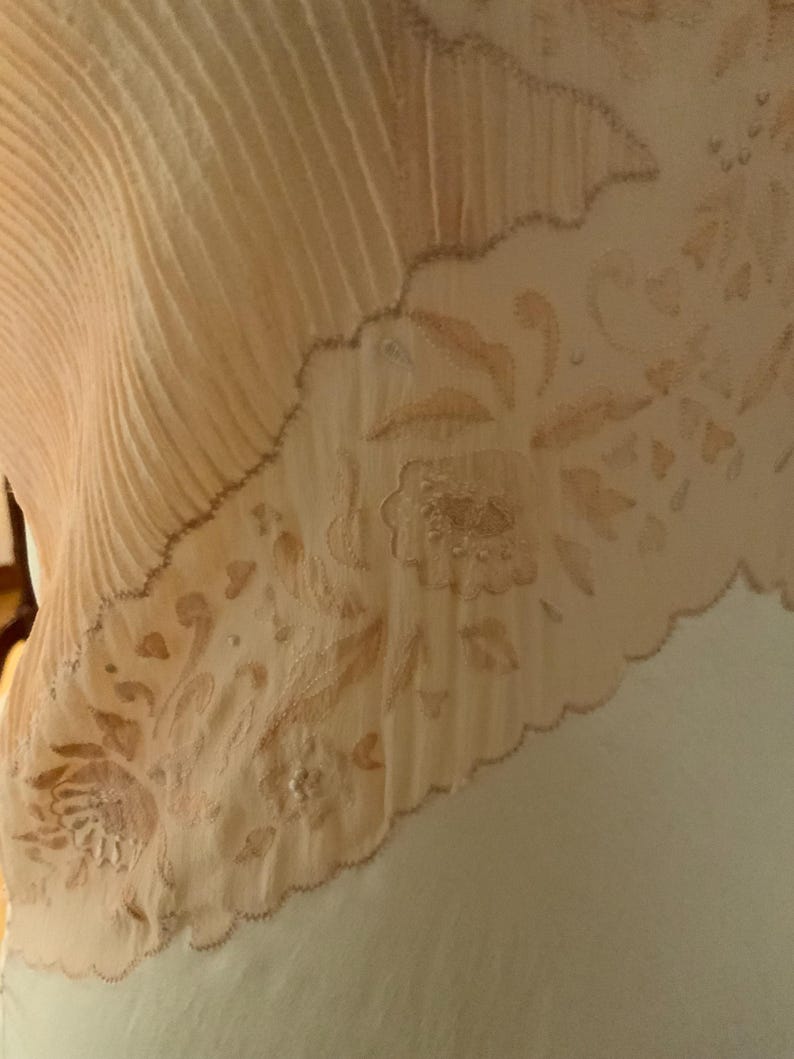 May include: Close-up of a cream-colored textile with a pleated section and a floral embroidered border. The embroidery features detailed flowers and leaves in a soft, muted peach color. The edge has a scalloped design.
