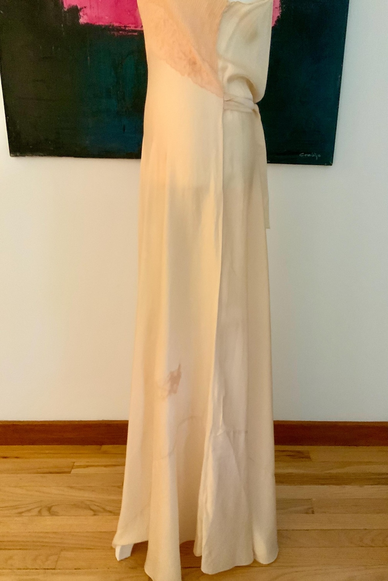 May include: A floor-length, cream-colored dress with a lace neckline. The dress has a flowing design and a subtle floral pattern near the hem. The dress is made of a soft, lightweight fabric.