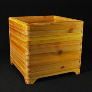 May include: A square wooden planter box constructed from light brown cedar planks with visible wood grain and knots. The sides are joined with interlocking finger joints at each corner, and the base features four small arched feet for elevation.