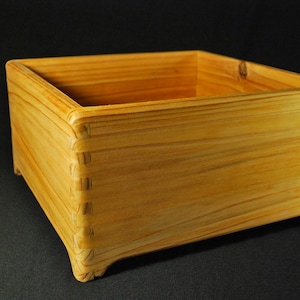 May include: A rectangular wooden box with a natural light brown finish and visible wood grain. The box features interlocking finger joints at each corner and four small, integrated feet at the base, providing a stable and open storage container.