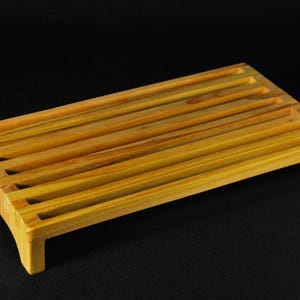 May include: A rectangular wooden bread slicing board featuring a series of parallel slats across the top surface. The board is constructed from light brown wood with a visible grain pattern and is supported by four short, sturdy feet at each corner.