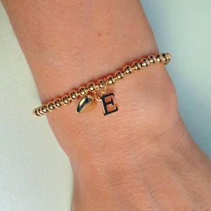 May include: Gold beaded bracelet with a heart charm and a black letter "E" charm. The bracelet is worn on a wrist and has a delicate, elegant appearance. The beads are small and evenly spaced.