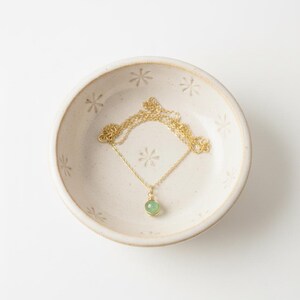 May include: A gold-colored necklace with a small, round, light green gemstone pendant rests in a cream-colored ceramic bowl. The bowl has a floral pattern and is set against a white background. The necklace chain is delicate.