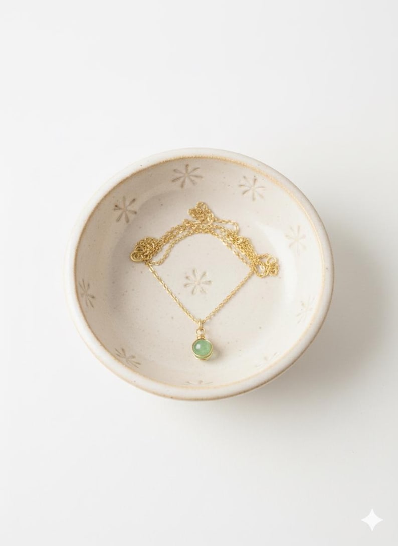 May include: A gold-colored necklace with a small, round, light green gemstone pendant rests in a cream-colored ceramic bowl. The bowl has a floral pattern and is set against a white background. The necklace chain is delicate.