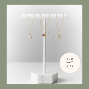 May include: A white marble jewelry display stand holding three gold necklaces with gemstone pendants. The pendants include a clear stone, a red stone, and a green stone. The stand has a hexagonal base and a circular graphic that says "YOU MAY LIKE".