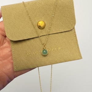 May include: A gold-colored necklace with a small, round, green gemstone pendant. The necklace is displayed on a tan pouch with the text "OLIVE & CLAY". The pouch has a gold-colored button closure.