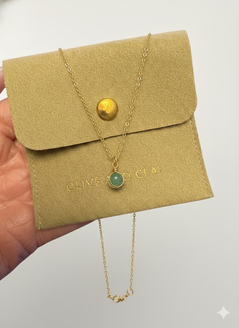 May include: A gold-colored necklace with a small, round, green gemstone pendant. The necklace is displayed on a tan pouch with the text "OLIVE & CLAY". The pouch has a gold-colored button closure.