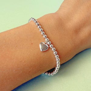 May include: A silver beaded bracelet with a heart charm. The bracelet is made of small, round beads and features a polished heart pendant. The jewelry is worn on a wrist.