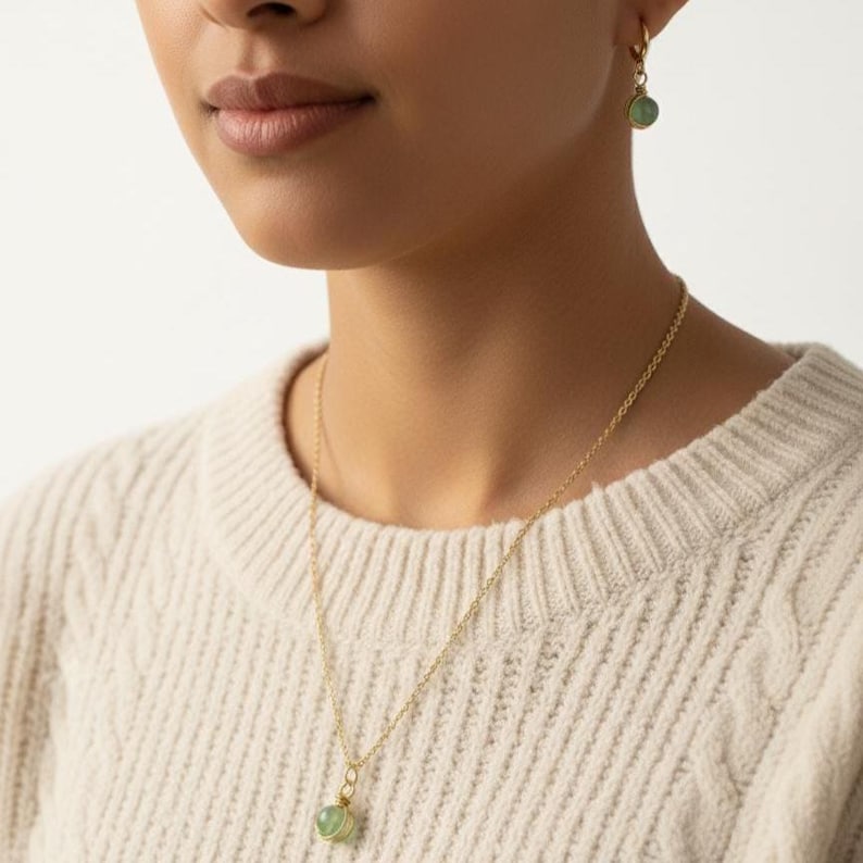 May include: A gold-colored necklace and matching earrings featuring a round, light green gemstone pendant and earring charms. The jewelry is worn on a person wearing a cream-colored cable knit sweater.