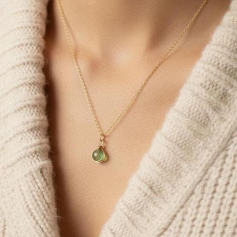 May include: A delicate gold necklace with a small, round, light green pendant. The pendant is suspended from a thin gold chain. The necklace is worn against a cream-colored sweater.