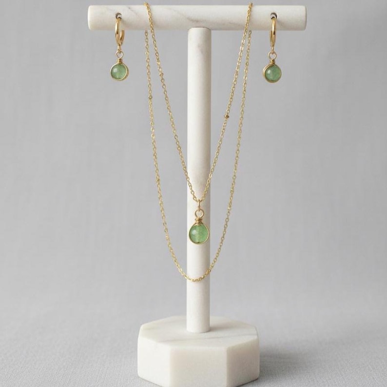 May include: A gold-colored jewelry set displayed on a white stand. The set includes a necklace with a green pendant and a pair of hoop earrings with matching green pendants. The stand has a hexagonal base.