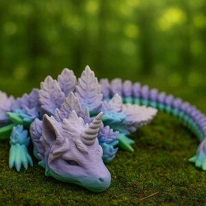 Könnte beinhalten: Eine Fantasy-Kreaturenfigur in Lila-, Blau- und Grüntönen, die einem Drachen oder Einhorn ähnelt. Die detaillierte Skulptur weist ein Horn, blattartige Stacheln und einen langen, segmentierten Schwanz auf. Die Figur liegt auf einem Bett aus grünem Moos.