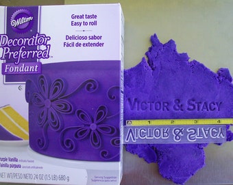 May include: A box of Wilton Decorator Preferred fondant next to a piece of purple fondant with the names "Victor & Stacy" imprinted. The fondant is measured with a ruler, approximately 5 inches wide. The box has text in English and Spanish.
