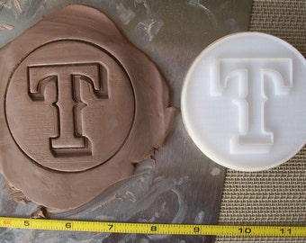 May include: A white plastic cookie cutter in the shape of the letter "T" with a decorative serif font. The cookie cutter is on a brown surface next to a brown clay imprint of the same letter.