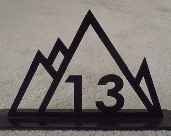 May include: A black metal mountain-shaped decoration with the number 13 cut out in the center. The design features three peaks and a rectangular base. The piece is likely used as a decorative item or a table centerpiece.