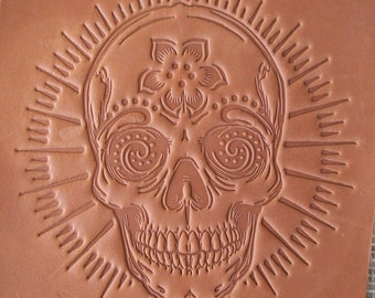 Può includere: Intaglio dettagliato in pelle marrone di un teschio di zucchero. Il teschio presenta intricati disegni floreali e a spirale, con linee radiali attorno alla testa. L'intaglio è centrato sulla pelle.