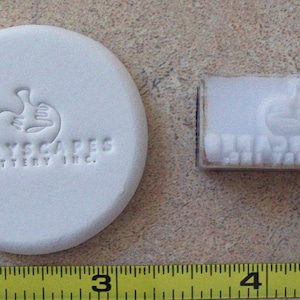 May include: White ceramic stamp with the text "Clay Scapes Pottery Inc." and a stylized hand design. A separate white ceramic stamp with the same hand design and the text "Clay Scapes Pottery Inc." is also shown.