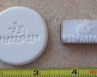 Può includere: Un timbro in ceramica bianca con il testo "Clay Scapes Pottery Inc." e un design a mano stilizzato. È mostrato anche un timbro separato in ceramica bianca con lo stesso design a mano e il testo "Clay Scapes Pottery Inc."