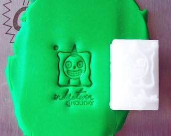 Può includere: Un timbro in polimero bianco con un personaggio dei cartoni animati che indossa un cappello da festa e il testo "indie twin holiday". Il timbro è premuto nell'argilla da modellare verde.