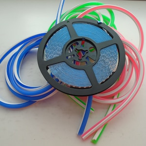 May include: A spool of blue LED light strip with a white and pink LED light strip wrapped around it. The spool is black and has a white label with the text "LED Strip Light".