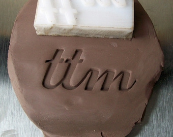 May include: A brown clay imprint of the word "time" with a white plastic stamp of the word "now" on top of it.