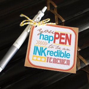 May include: A brown card with the text "you hapPEN to be an INKredible TEACHER" in colorful letters. Three yellow apple icons are below the text. A white pen with a yellow and white string is tied around it.