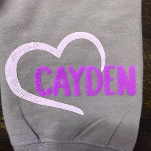 Op de afbeelding: Een grijs sweatshirt met een witte hartlijn en de naam "Cayden" in paarse letters binnen het hart.