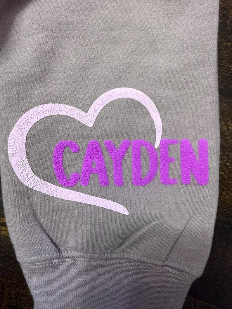 Op de afbeelding: Een grijs sweatshirt met een witte hartlijn en de naam "Cayden" in paarse letters binnen het hart.
