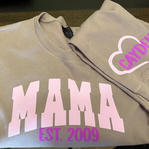 Op de afbeelding: Een mauve sweatshirt met het woord "MAMA" in roze letters en de tekst "EST. 2009" in paarse letters. Het sweatshirt heeft een bijpassende mauve mouw met een wit hart en het woord "CAYDEN" in paarse letters.
