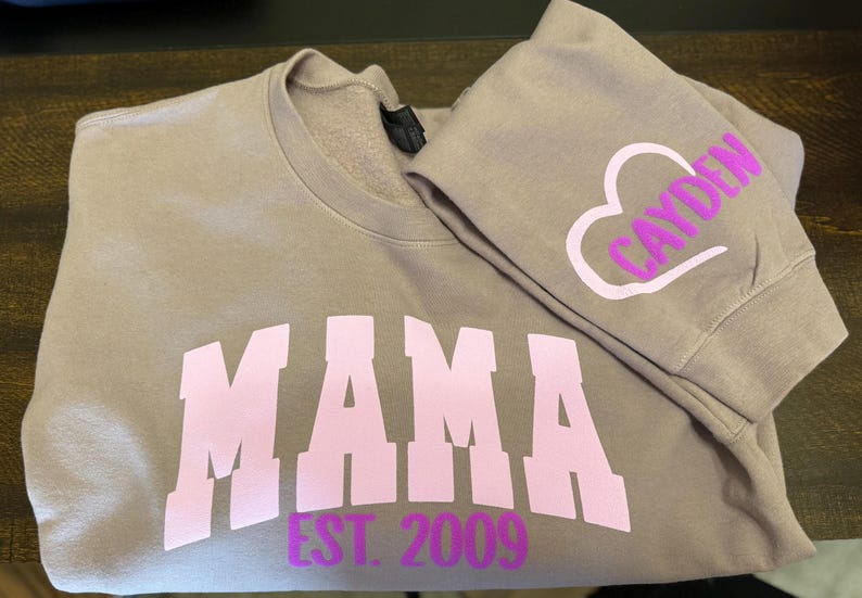 Op de afbeelding: Een mauve sweatshirt met het woord "MAMA" in roze letters en de tekst "EST. 2009" in paarse letters. Het sweatshirt heeft een bijpassende mauve mouw met een wit hart en het woord "CAYDEN" in paarse letters.