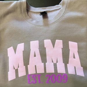 Op de afbeelding: Een grijs sweatshirt met het woord "MAMA" in roze letters en "EST. 2009" in paarse letters.