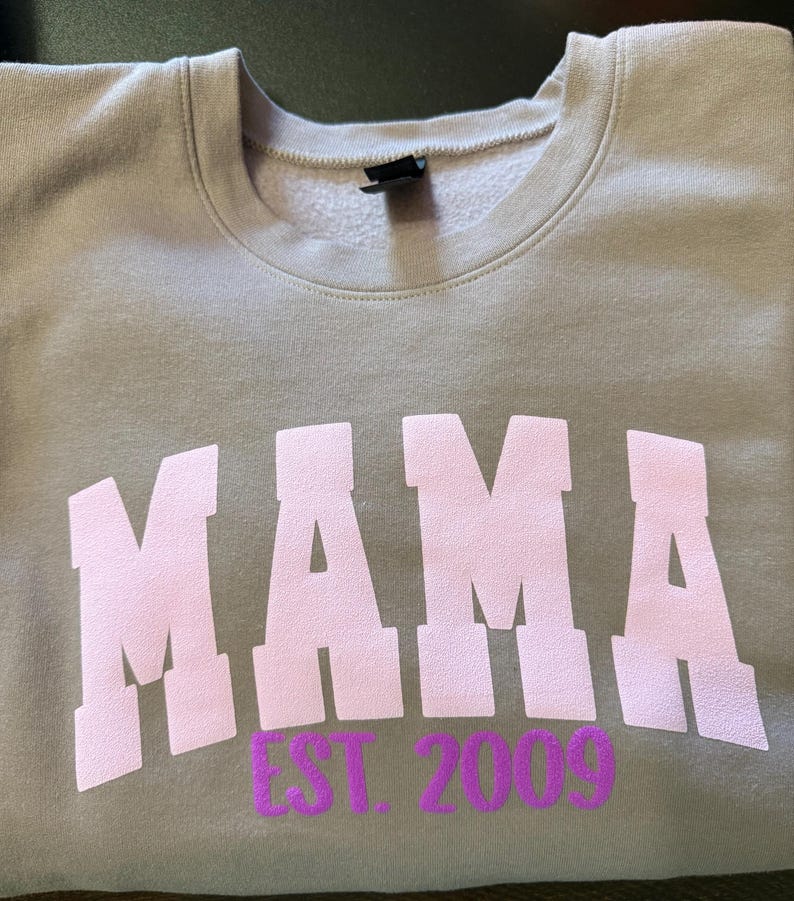 Op de afbeelding: Een grijs sweatshirt met het woord "MAMA" in roze letters en "EST. 2009" in paarse letters.