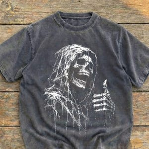 Op de afbeelding: Een antracietgrijs t-shirt met korte mouwen en een witte grafische print van een skelet met capuchon en een spinnenwebtextuur. Het skelet is afgebeeld met een open mond en een skeletachtige hand die een duim omhoog gebaar maakt op de voorkant.
