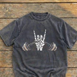 Peut inclure: T-shirt gris fonc&eacute; avec un motif de main de squelette blanche tenant une barre de musculation. Le design inclut le geste de la main rock and roll. Le t-shirt a un aspect vintage et us&eacute;. Le texte "Worn Heritage Vintage Wear" est en bas.