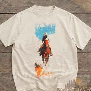 May include: Ivory t-shirt with a vintage graphic of a cowboy on horseback. The design includes a blue sky, flames, and the words "Worn Heritage Vintage Wear". The shirt is a classic crew neck.