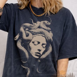 May include: Black comfort colors t-shirt featuring a Medusa graphic. The design depicts a woman's face with snakes for hair. The shirt has the text "Worn Heritage Vintage Wear" and "black comfort colors".