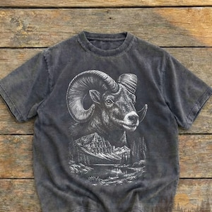 Könnte beinhalten: Anthrazitfarbenes T-Shirt mit weißer Illustration eines Widderkopfes und einer Berglandschaft. Das T-Shirt hat einen Vintage-Wascheffekt und den Text "Worn Heritage Vintage Wear". Das Design zeigt detaillierte Widderhörner.