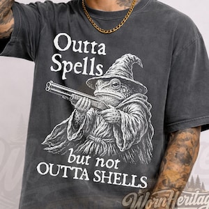 May include: A charcoal gray short-sleeve t-shirt featuring a white graphic of a toad wearing a wizard hat and holding a shotgun. The text reads Outta Spells but not OUTTA SHELLS. The shirt has a faded, vintage-style wash and a crew neckline.