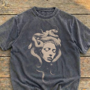 Op de afbeelding: Een antracietgrijs t-shirt met korte mouwen en een vervaagde, acid-wash uitstraling. Het midden van het shirt bevat een beige grafische illustratie van een menselijk gezicht met slangen verstrengeld in het haar, lijkend op de mythologische Medusa.