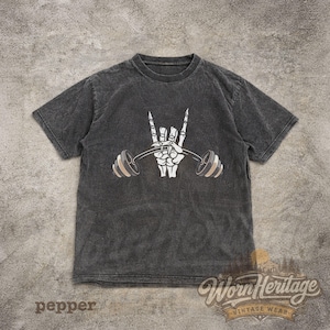 Peut inclure: T-shirt &agrave; manches courtes de couleur poivre avec une main de squelette blanche soulevant une barre. Le motif comprend un geste de la main rock and roll. Le t-shirt a un lavage vintage et le texte "Worn Heritage Vintage Wear".