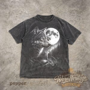 Op de afbeelding: Een donkergrijs t-shirt met een gedetailleerde illustratie van een wolf die naar de maan huilt. Het ontwerp bevat een grote wolfskop, een volle maan en een kleinere wolf in een bosrijke omgeving. De tekst "Worn Heritage Vintage Wear" is zichtbaar.