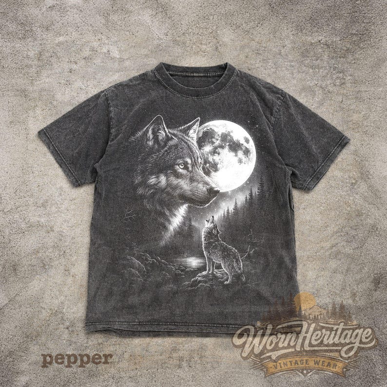 Op de afbeelding: Een donkergrijs t-shirt met een gedetailleerde illustratie van een wolf die naar de maan huilt. Het ontwerp bevat een grote wolfskop, een volle maan en een kleinere wolf in een bosrijke omgeving. De tekst "Worn Heritage Vintage Wear" is zichtbaar.